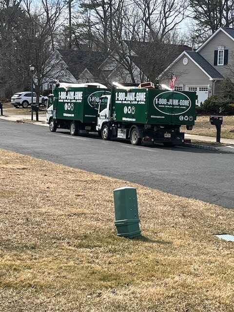 Two green junk removal trucks parked on a residential street, with a house and mailbox visible in the background.