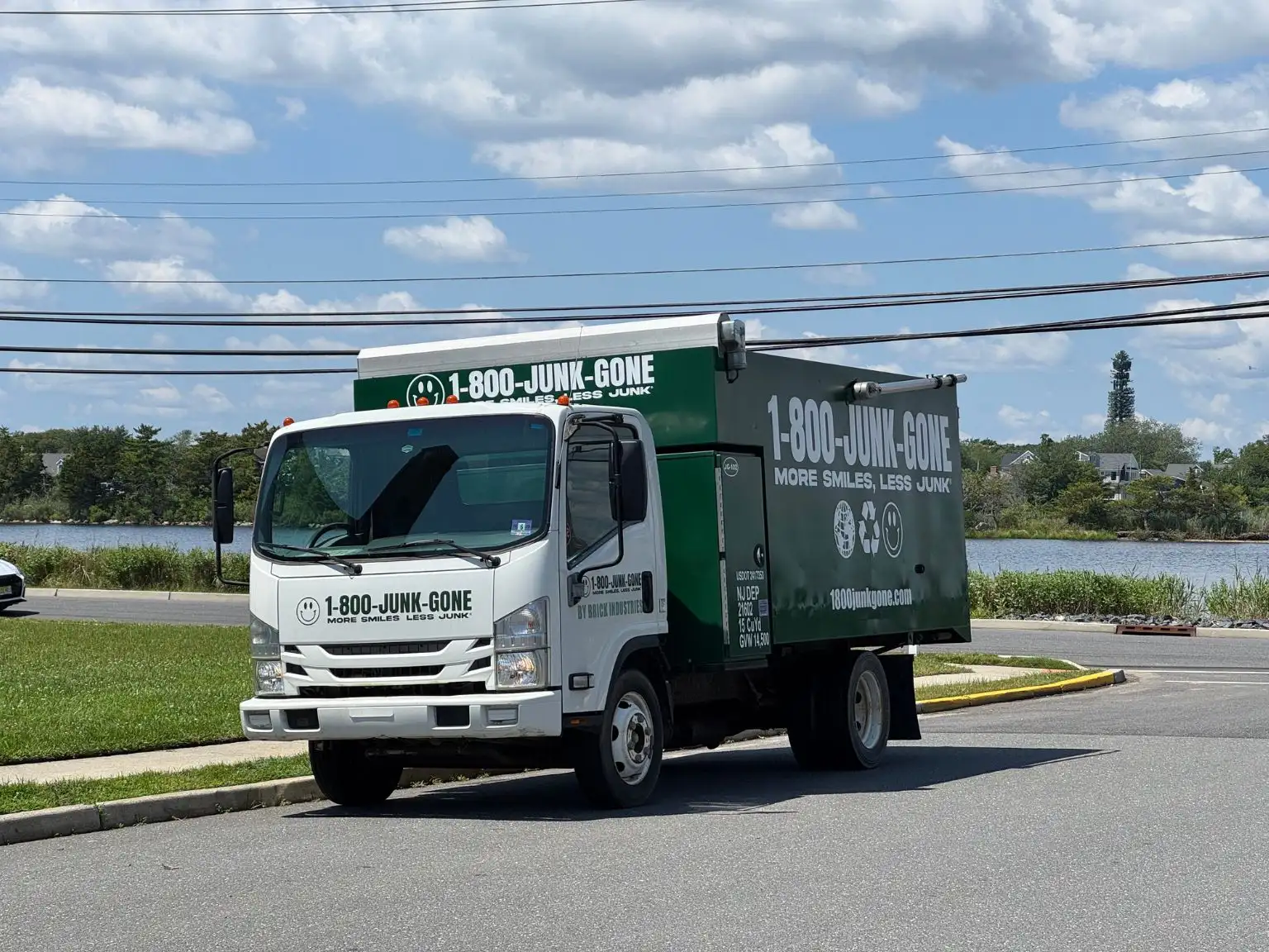 Three-quarter view of a white and green 1-800-JUNK-GONE truck parked on an asphalt street near water under a cloudy sky