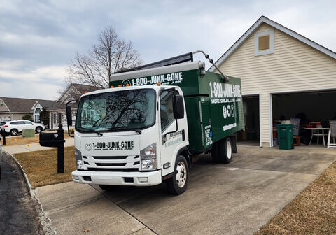 A green junk removal truck parked in a driveway beside a residential garage on a cloudy day.