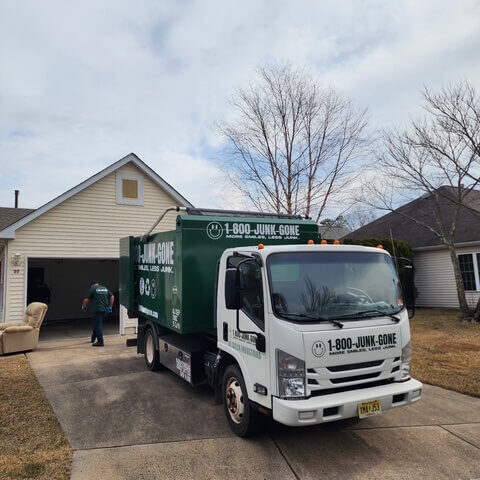 A green 1-800-JUNK-GONE truck parked in front of a house, with a worker collecting junk on a cloudy day.