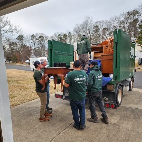 Three-movers-in-green-shirts A group of movers in green shirts loading wooden furniture into the back of a truck on a cloudy day.