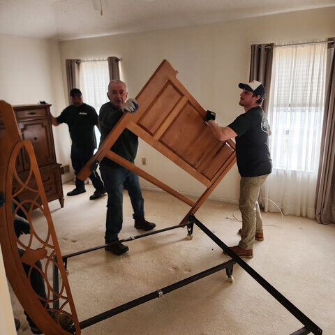 Two-men-are-lifting-a-large-piece-of-furniture Two men lift a large wooden piece of furniture while a third man assists, in a bright room with neutral walls and curtains.