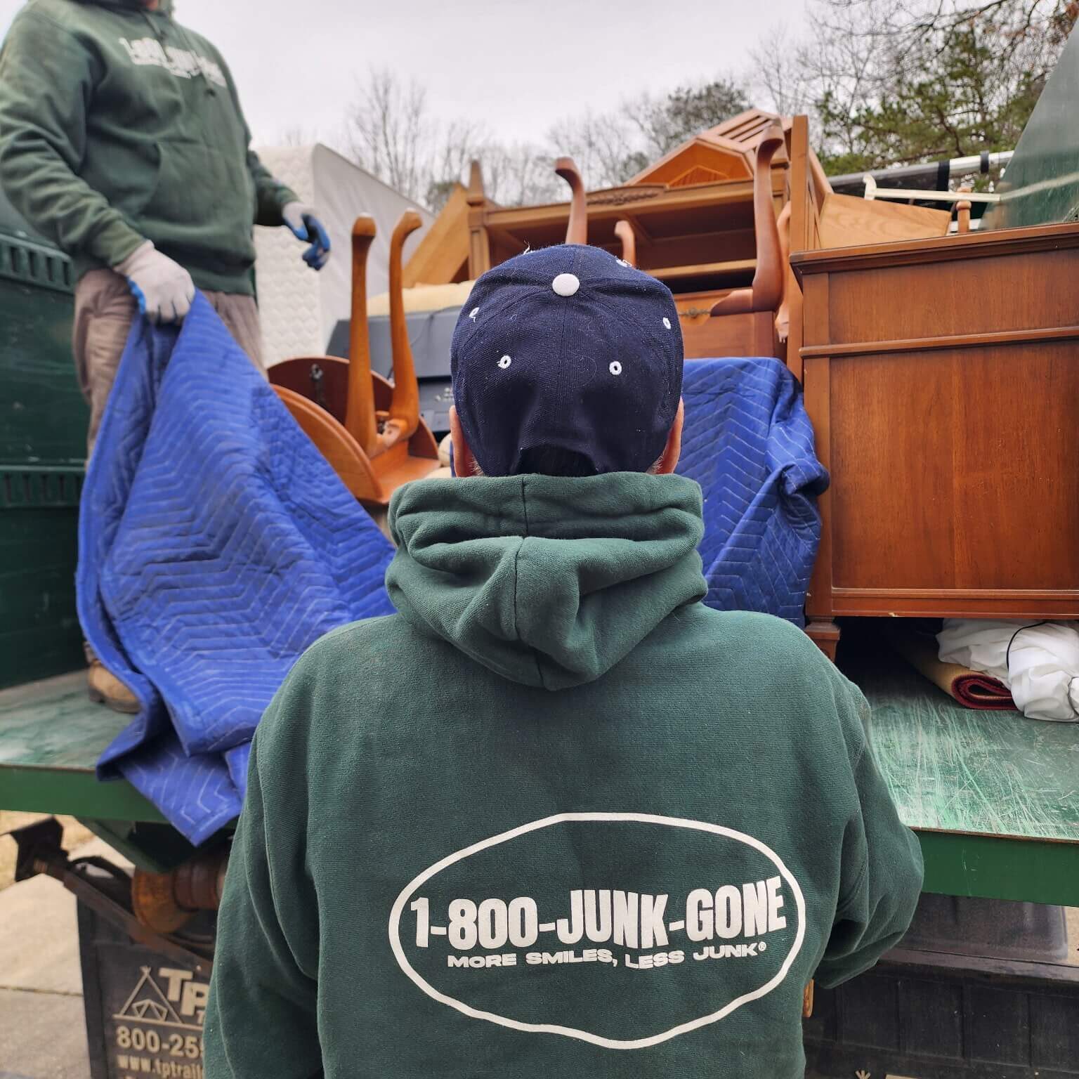 Two-workers-in-green-sweatshirts Two workers in green hoodies load furniture onto a truck covered with a blue blanket, preparing for junk removal.