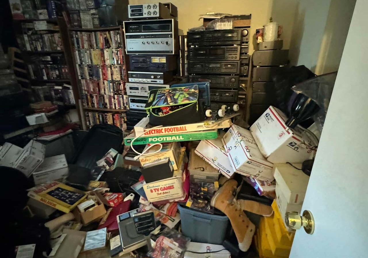 A cluttered room filled with various electronics and stacks of books, showcasing disorganization and a busy atmosphere.
