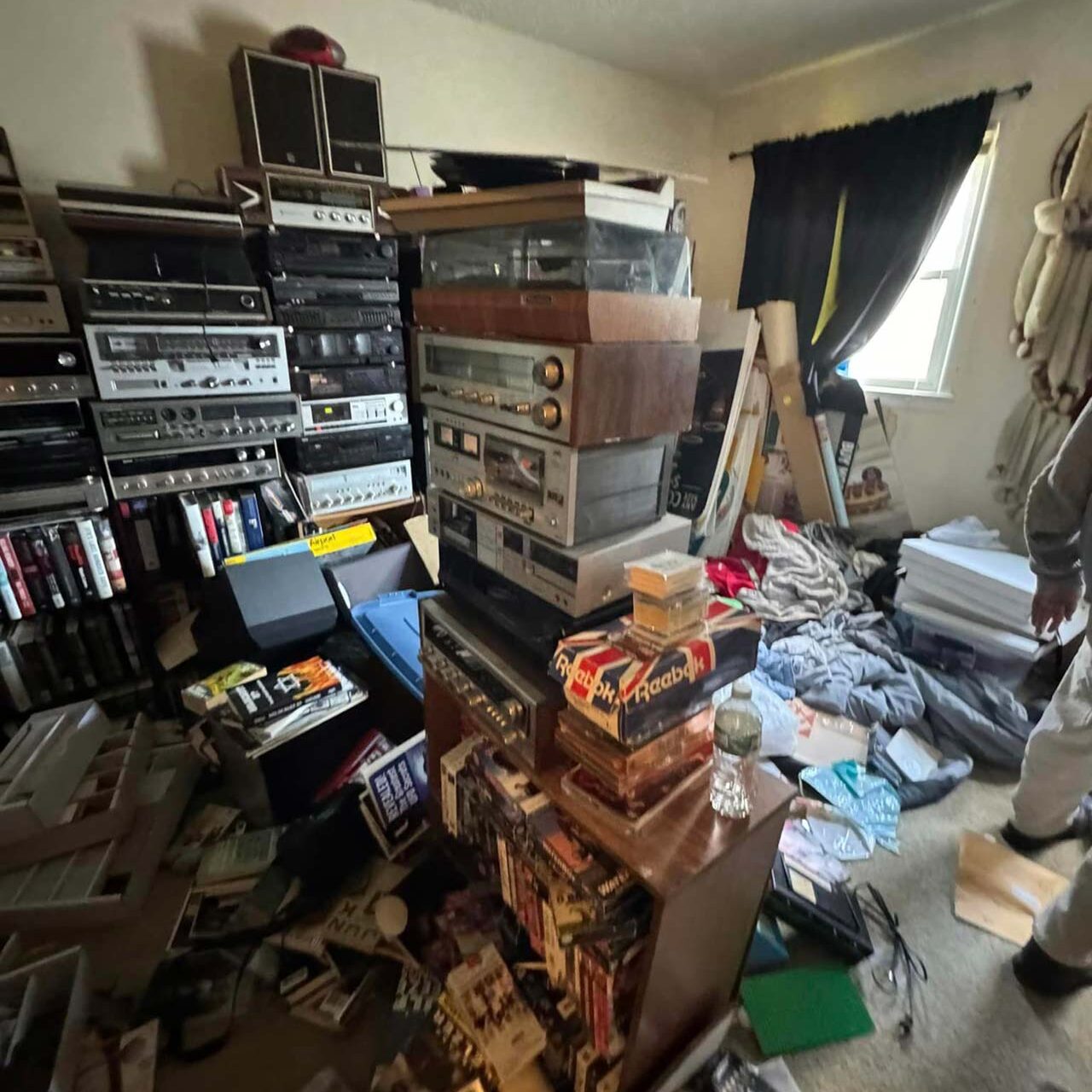 A man stands in a cluttered room filled with various items and debris, highlighting disorganization and chaos.