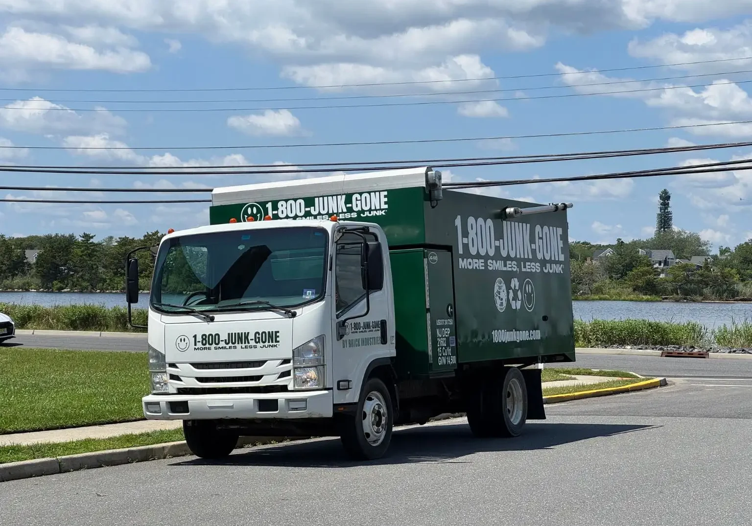 Three-quarter view of a white and green 1-800-JUNK-GONE truck parked on an asphalt street near water under a cloudy sky