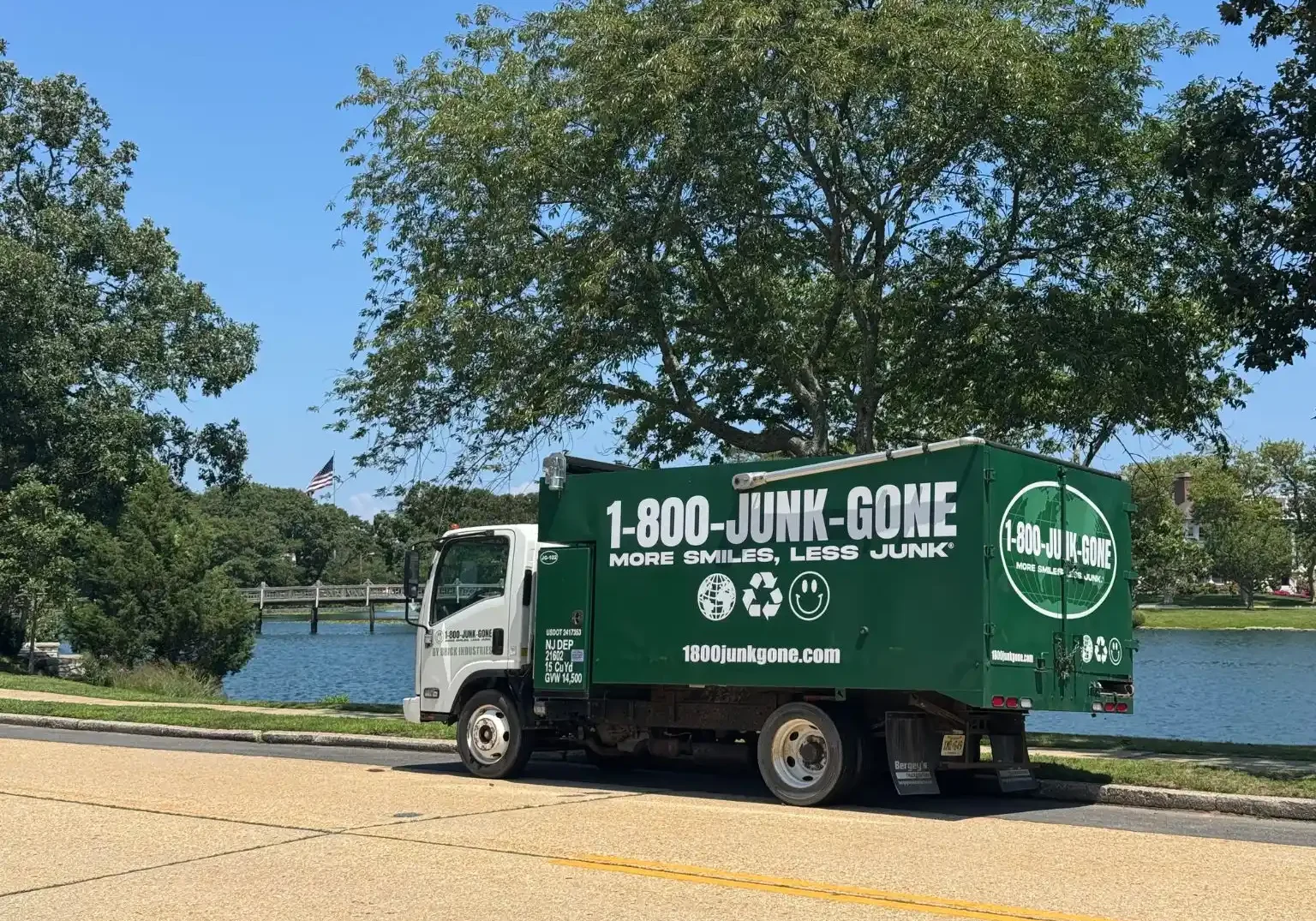 Full side profile of a green 1-800-JUNK-GONE junk removal truck parked along a waterfront road beneath a large tree
