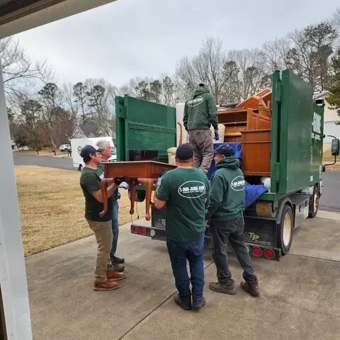 A group of movers in green shirts loading wooden furniture into the back of a truck on a cloudy day.